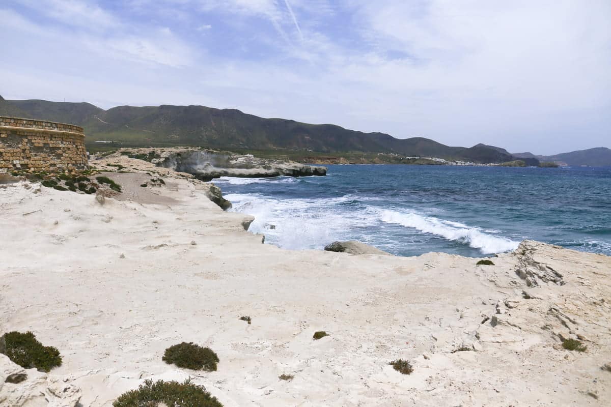 Rocky white coastline with sparse vegetation, blue ocean waves crashing against the shore, and distant green hills under a partly cloudy sky. Escullos Beach in Cabo de Gata, Almería
