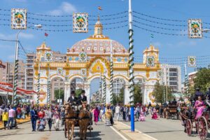 horse carriages with people dressed in traditional Andalusian costumes and a large arc at the end of the street; Feria de Abril in Seville