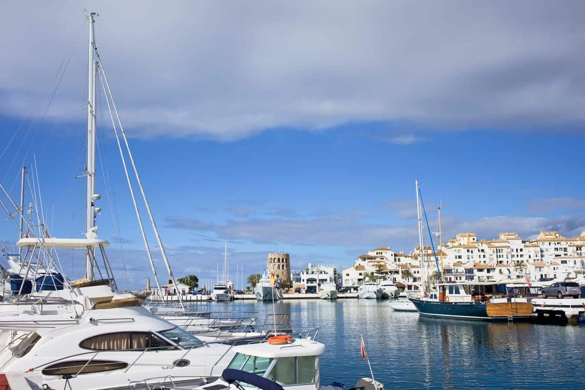 a marina with luxurious yachts and at the background white buildings and a rounded tower; the Marina of Puerto Banus near Marbella