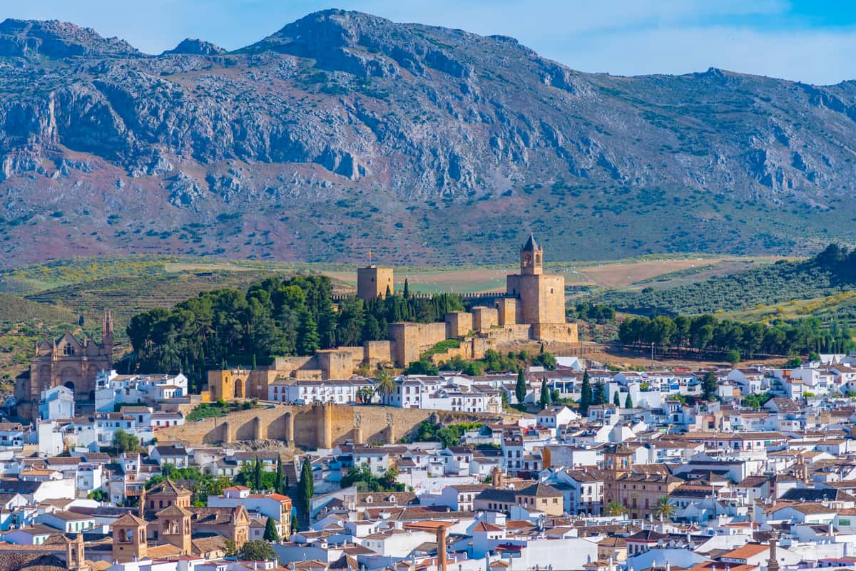 A picturesque town is nestled at the base of a rugged mountain range. Central to the image is Antequera's historic castle with stone walls and towers, surrounded by lush greenery. Whitewashed buildings with tiled roofs populate the foreground under a clear blue sky, reflecting timeless charm.
