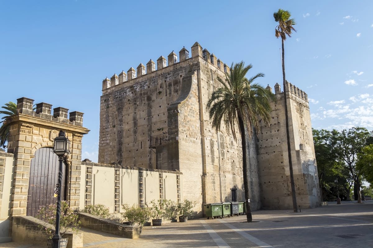 A stone fortress with crenellated battlements stands under a clear blue sky. The historic Alcazar of Jerez, featuring a rectangular tower and palm trees in the foreground, is surrounded by a paved courtyard with a lamppost and some greenery.