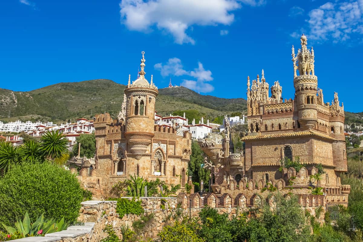 A stone castle features intricate architectural details against a backdrop of green hills on a clear, sunny day. The bright blue sky is dotted with a few white clouds. Vegetation surrounds the castle, and white buildings are visible in the background. Castillo de Colomares in Benalmadena