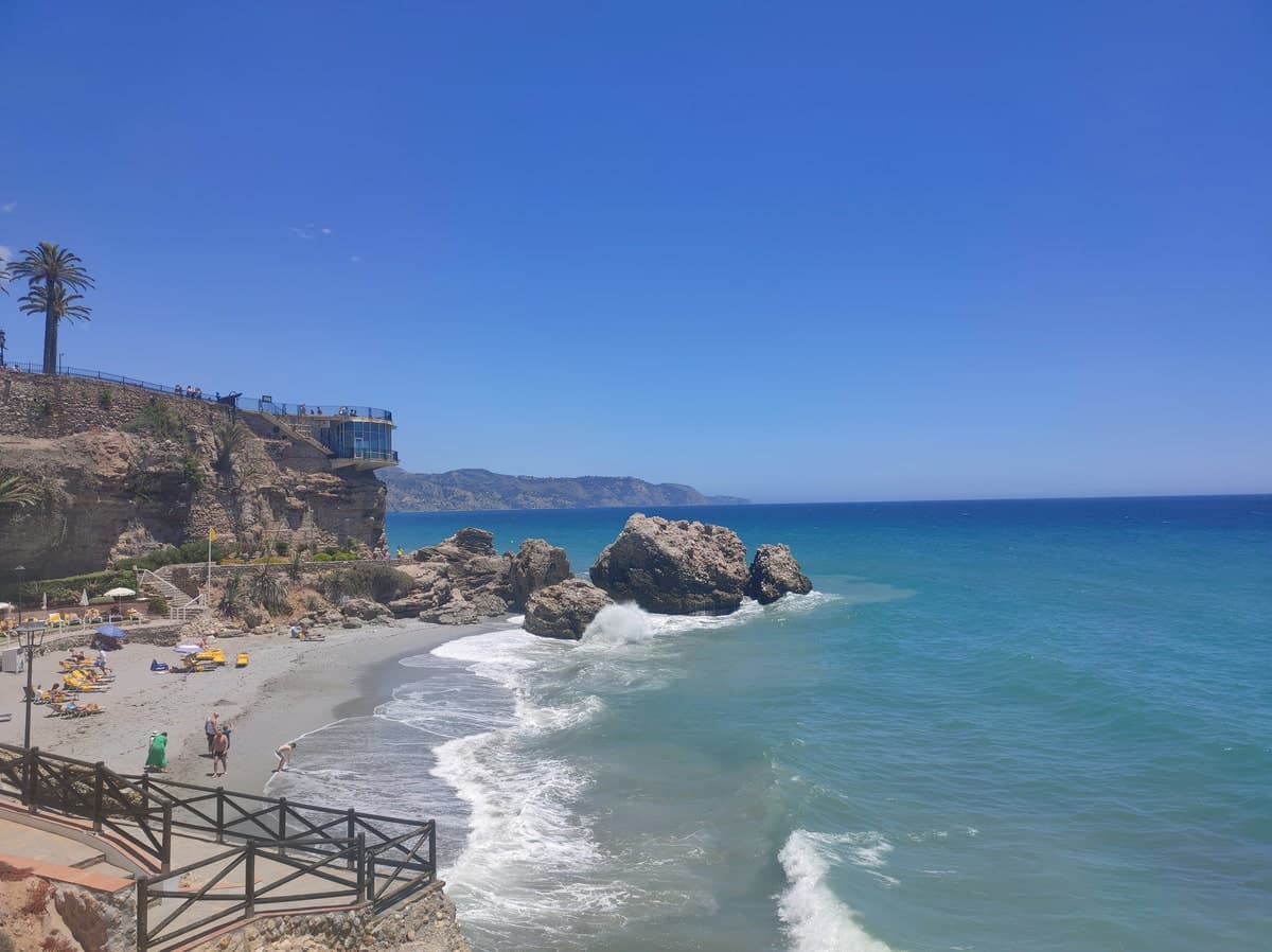 A coastal scene featuring a sandy beach with a few people and scattered sunbeds. Rocky cliffs outline the shore to the left, topped with palm trees and a patio. Clear blue sky and turquoise sea waves characterize this seaside view. El balcon de Europa in Nerja