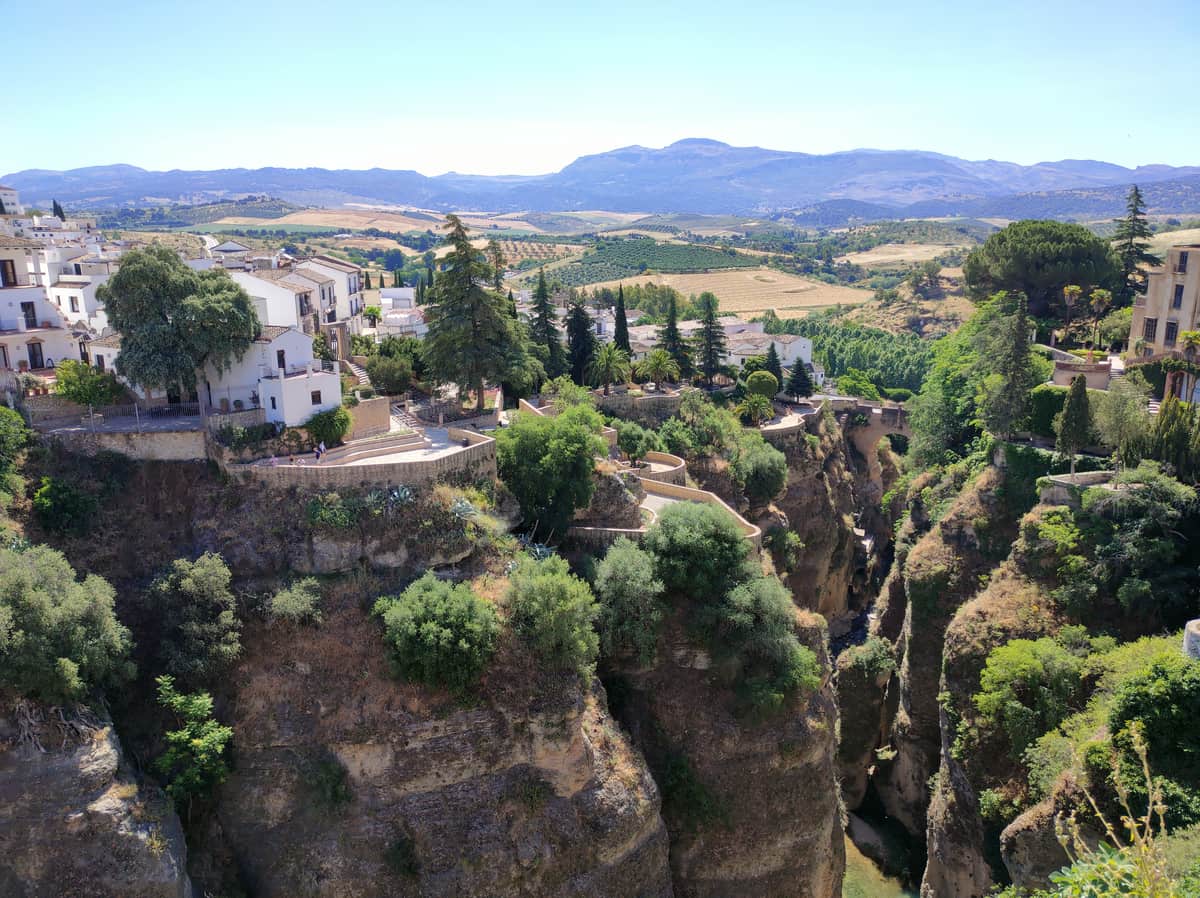 A scenic view of Ronda, Spain, showcasing white buildings perched on the edge of a deep gorge. Trees and greenery are scattered throughout the area, with mountains visible in the background, under a clear blue sky.