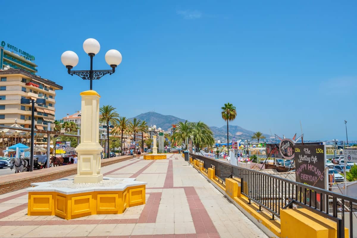 A seaside promenade on a clear day, featuring a lamp post with three spherical lights. The walkway is lined with palm trees and bordered by a railing on one side. Buildings, including a hotel, and mountains are visible in the background. Fuengirola Promenade