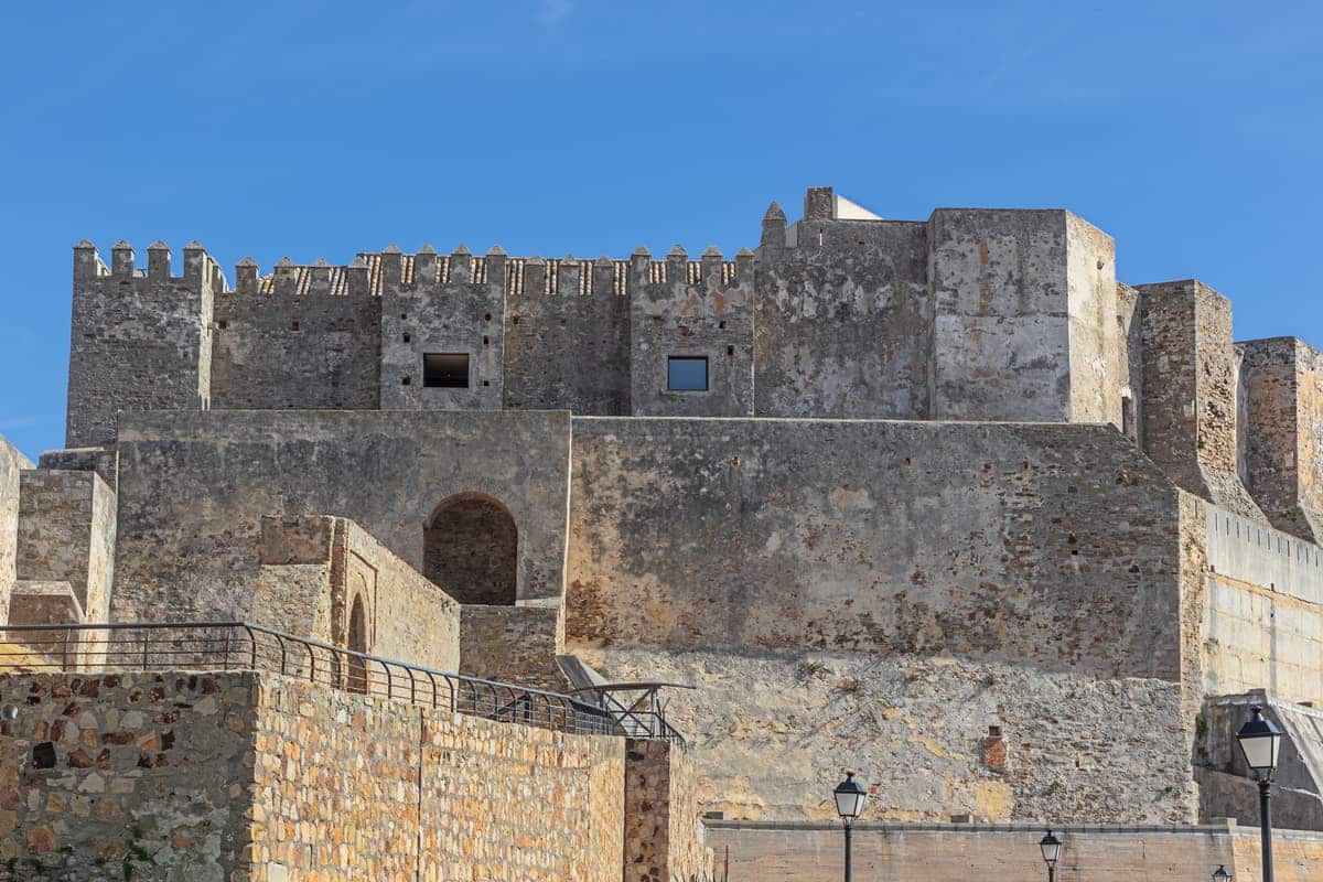 A view of an old stone fortress with multiple levels, battlements, and watchtowers under a clear blue sky. The weathered stone walls exhibit various shades of gray and beige. A few street lamps are positioned near the base of the walls. Guzman el Bueno Castle in Tarifa