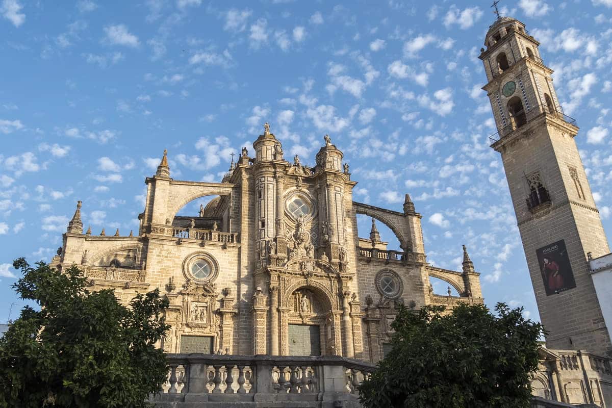 A large historic cathedral made of stone with intricate architectural details and multiple towers under a blue sky with scattered clouds. In the foreground are green trees, and to the right, a tall bell tower stands with a visible clock. Jerez de la Frontera Cathedral