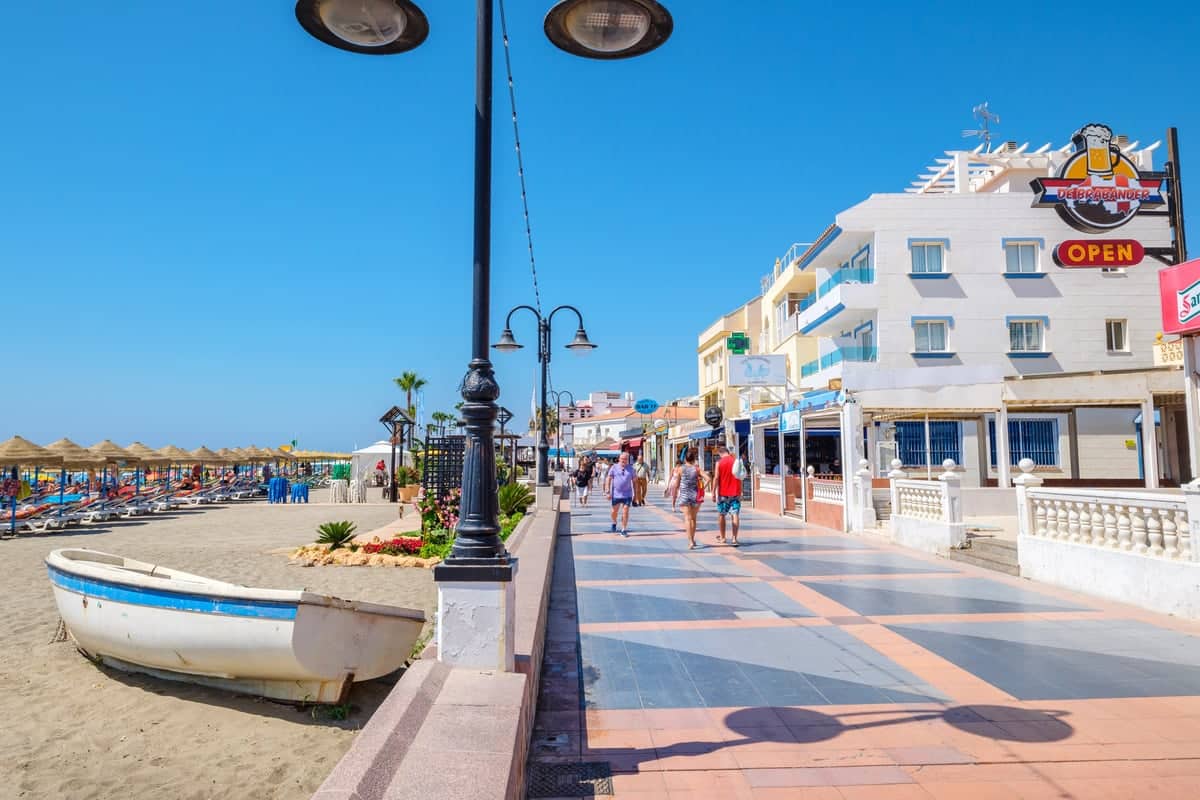 A sunny day at a beachfront promenade featuring a row of shops and restaurants on the right, beach umbrellas and lounge chairs on the left, and several people walking along the walkway. A small boat is positioned on the sand near the promenade. La Carihuela promenade in Torremolinos