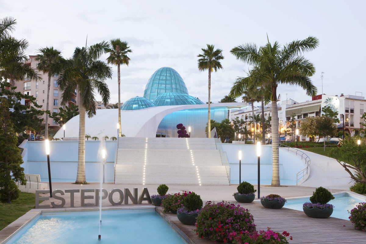 An urban park features a modern, white, wave-shaped building with a blue-tinted glass dome. Palm trees, street lamps, and planters with flowers are in the foreground. A minor pool with a sign reading "ESTEPONA" is visible at the image's bottom left corner. The Orchidarium in Estepona, Spain