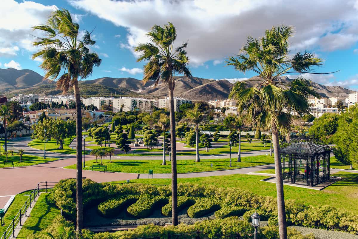 A landscaped park with palm trees, green lawns, and neatly trimmed shrubs. A gazebo-like structure is on the right. In the background, there are residential buildings and mountainous terrain under a partly cloudy sky. Paths crisscross the park area. Parque la Batería in Torremolinos