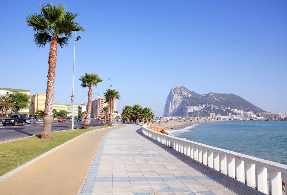 A seaside promenade with palm trees on the left and a railing on the right overlooking the water. In the background, there is a prominent rock formation known as the Rock of Gibraltar, with city buildings in the vicinity. Clear blue sky overhead. Playa de Poniente in La Línea, Andalusia