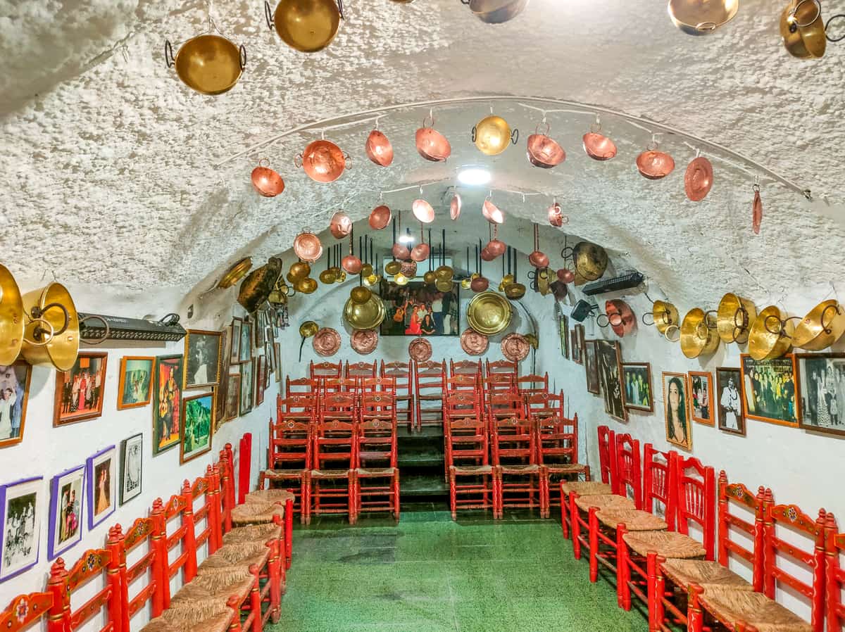 a cave with a rounded ceiling, two rows of seats at the sides and a few seats at the back with photographs of flamenco dancers hanging on the walls and copper and copper and messing cookware hanging from the roof and on the walls; a flamenco cave in Sacromonte, Granada
