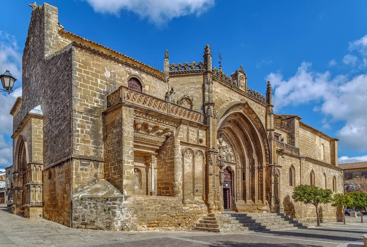 A historic stone cathedral with detailed carvings above the main entrance and a mix of Gothic and Romanesque architectural elements. The building features arched windows, a grand staircase, and is situated on a paved plaza under a clear blue sky. San Pablo Church in Úbeda