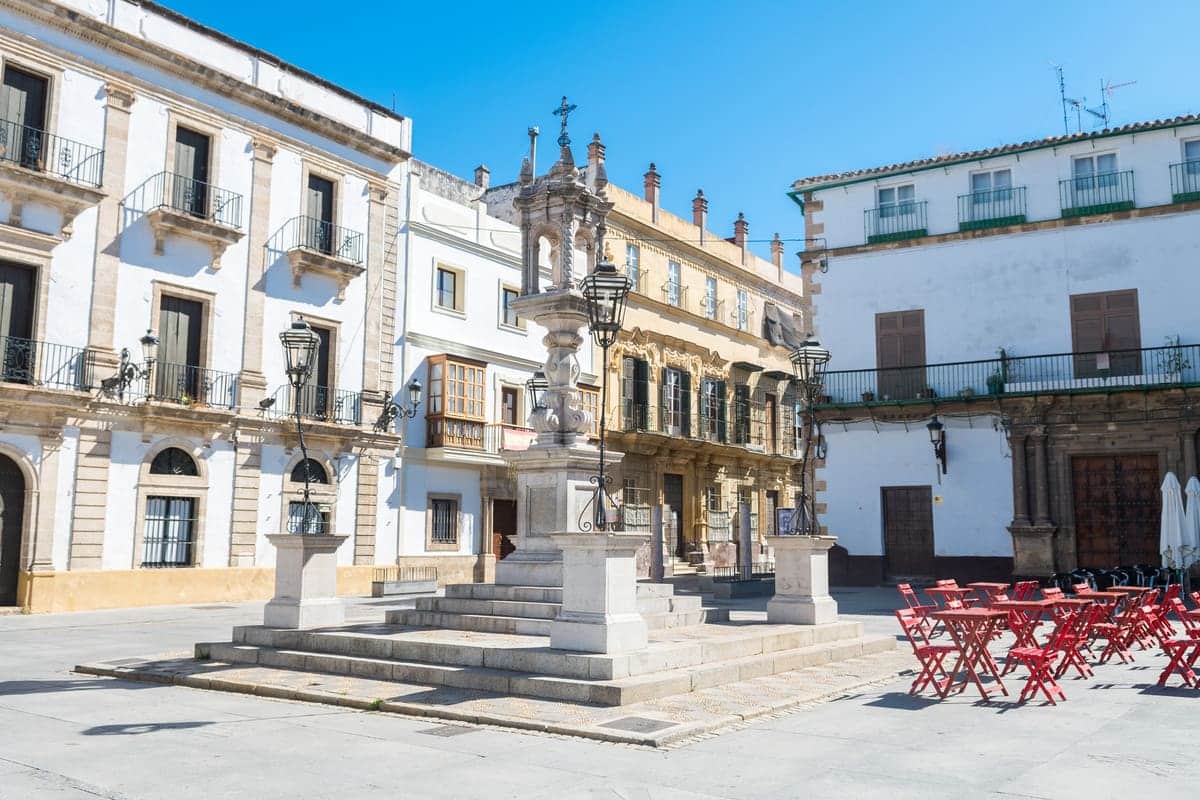 A sunny plaza with an ornamental fountain at the center, surrounded by historic multi-story buildings with balconies. The square has red tables and chairs set up on the right side near a white building. The clear sky is bright blue. Sanlúcar de Barrameda in Andalusia