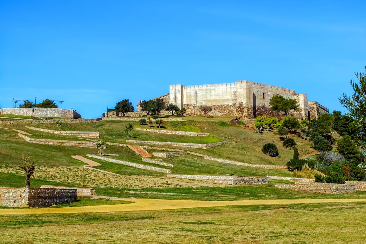 A historic stone fortress stands atop a grassy hill adorned with terraced landscaping under a clear blue sky. Pathways and staircases wind up the hill toward the fortress, with young trees and shrubs scattered throughout the scene. Sohail Castle in Fuengirola