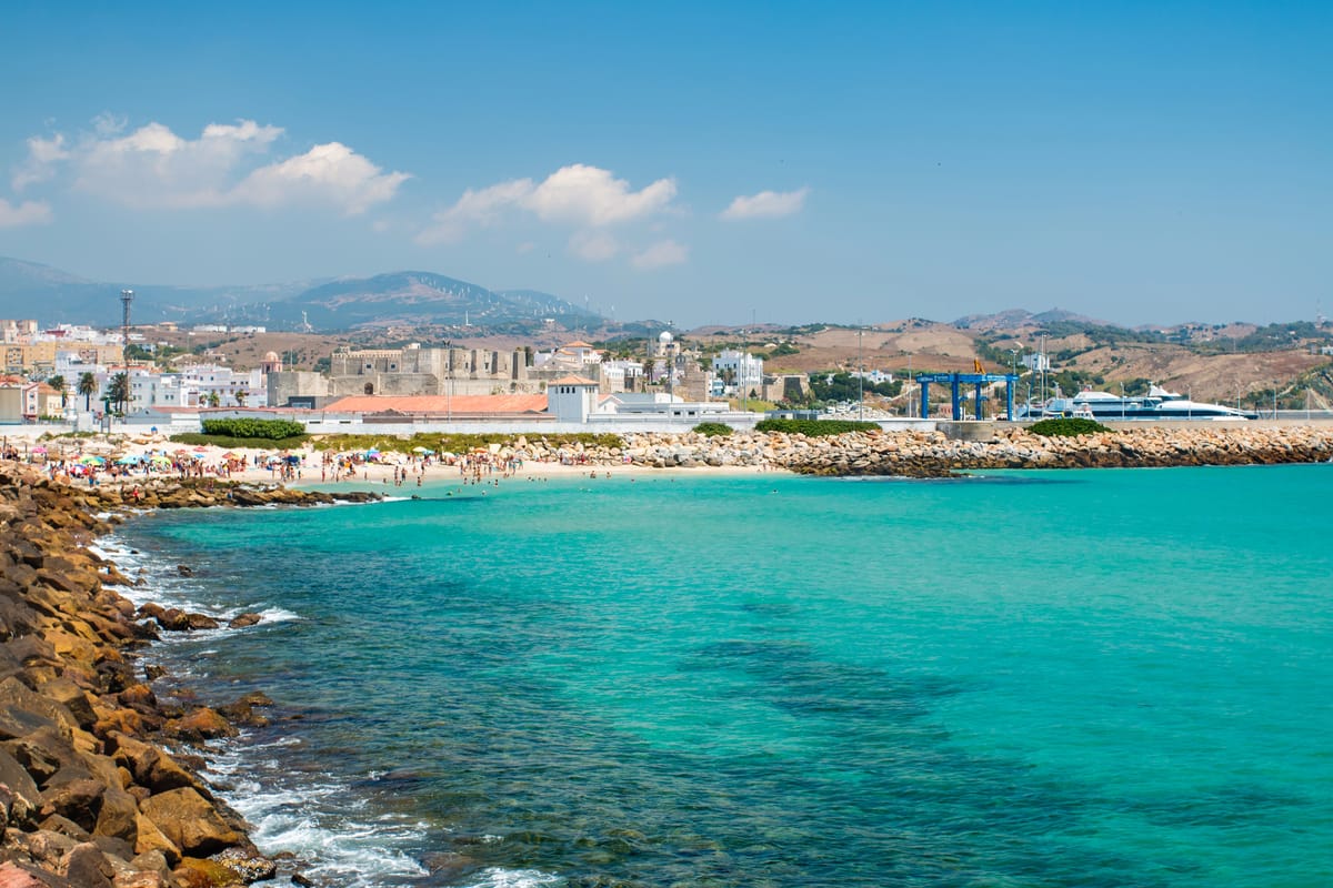A coastal scene featuring a sandy beach with people sunbathing and swimming. The foreground includes a rocky shoreline with clear turquoise water. In the background, there is a cityscape with buildings and mountains under a mostly clear blue sky. Tarifa in Andalusia