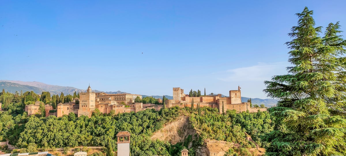 a view to a reddish-looking fortress on a green hill, with mountain tops at the back, and red rooftops in front; a view the Alhambra from Mirador de San Nicolas in Granada