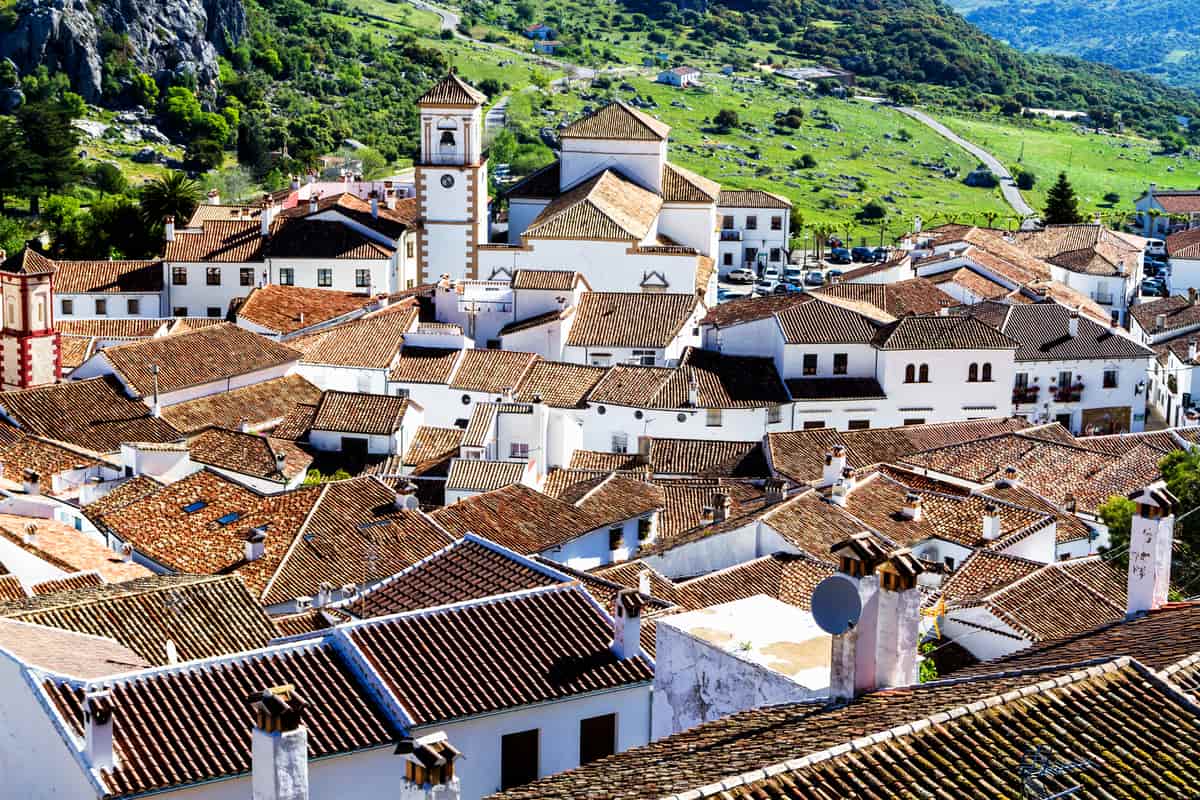 Scenic hilltop town featuring white buildings with terracotta roofs clustered around a central stone church with a bell tower and clock. The village is nestled among lush green hills and rocky cliffs, with a dramatic mountain backdrop under a partly cloudy sky. Grazalema pueblo blanco in Andalasia