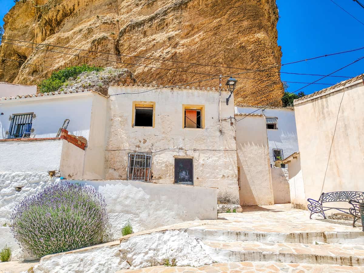White stucco houses with barred windows sit at the base of a large tan rock cliff under a clear blue sky; a lavender bush, stone steps, and a metal bench are visible in the foreground. Barrio del Arrabal in Montefrío