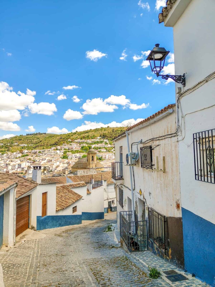 A narrow cobblestone street lined with whitewashed buildings and wrought iron window grilles descends toward a town with tiled rooftops, with green hills and scattered clouds visible in the background. A street in Montefrío