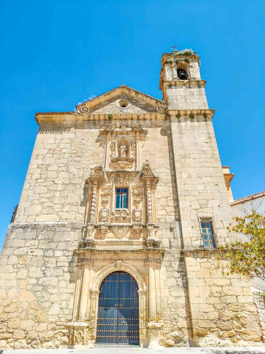 A historic stone church with ornate carvings above the arched entrance, a single tower on the right side, and a blue sky in the background. The Church of San Antonio in Montefrío