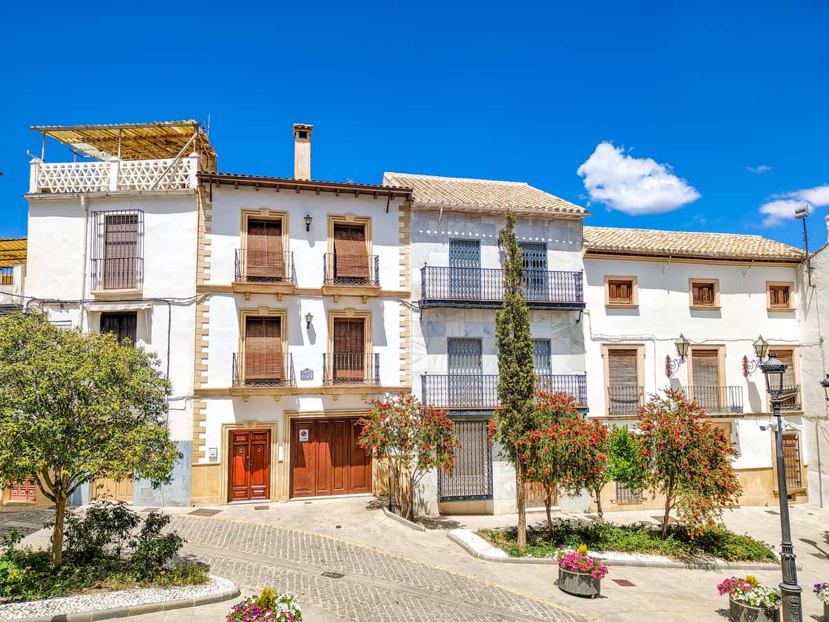 A sunny street scene shows traditional white buildings with tile roofs, balconies, and shuttered windows. Potted plants, small trees, and flowering bushes line the cobblestone pavement under a clear blue sky. Houses at Plaza de España, Montefrío