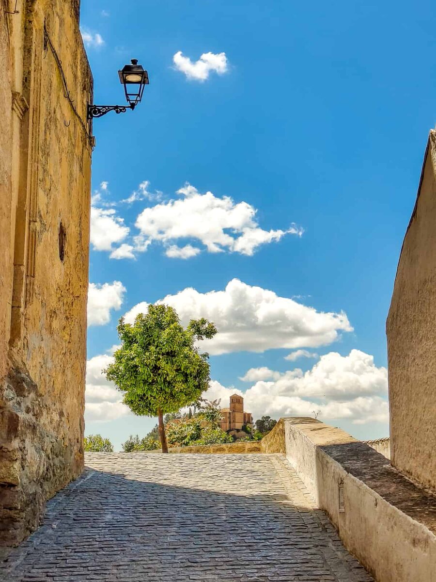 A narrow cobblestone street curves uphill between old stucco walls, with a tree and a distant historic building under a blue sky with scattered clouds. A black street lamp is mounted on the left wall. Mirrador Placeta de Convento in Montefrío