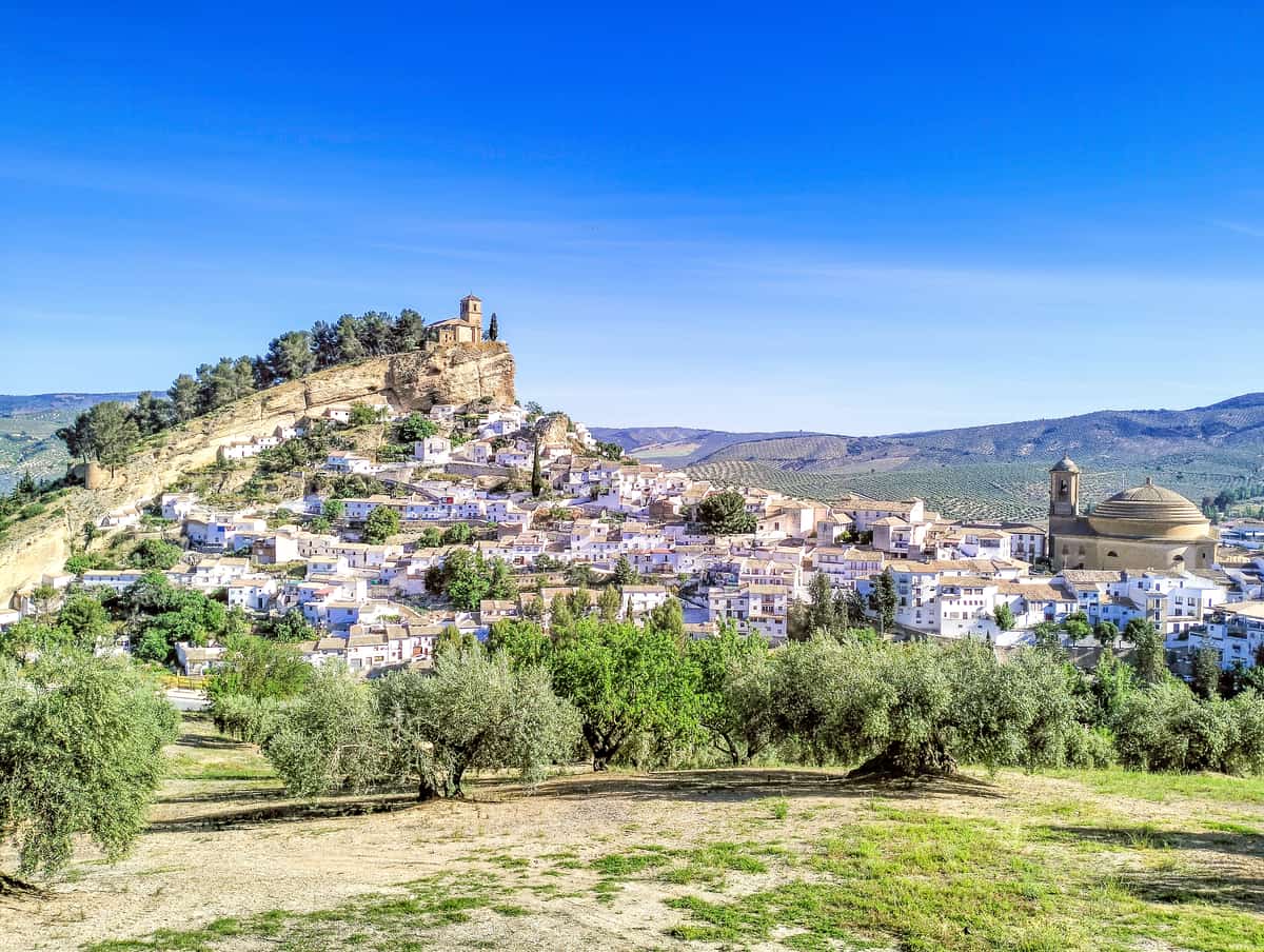 A hillside town with white buildings, a church on top of the hill, a domed building in the foreground, and olive trees in the foreground under a clear blue sky. Montefrío Granada, one of the most beautiful villages in Spain