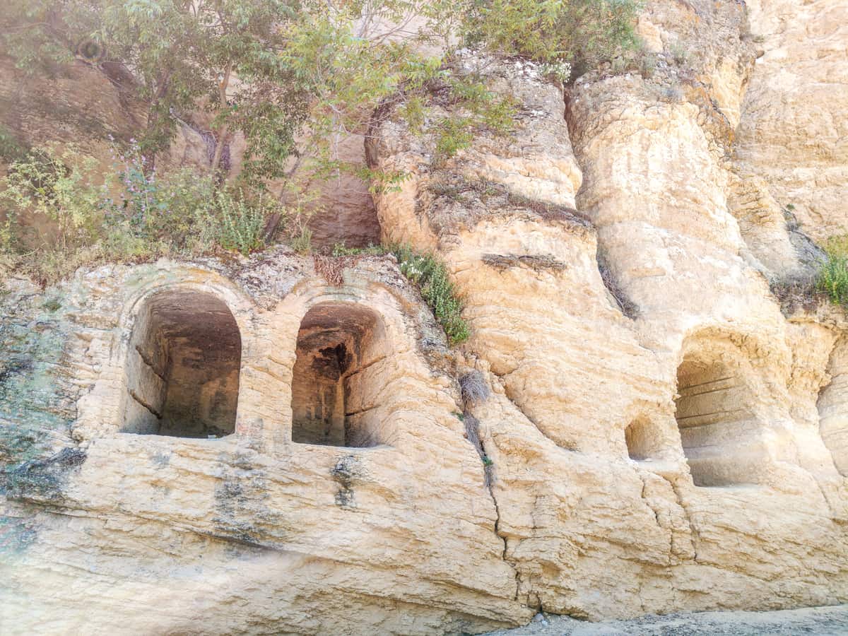 Rock formations with three arched openings carved into the cliffside, surrounded by sparse vegetation and sunlight filtering through the scene. The Pantheon - Old Graveyard in Montefrío
