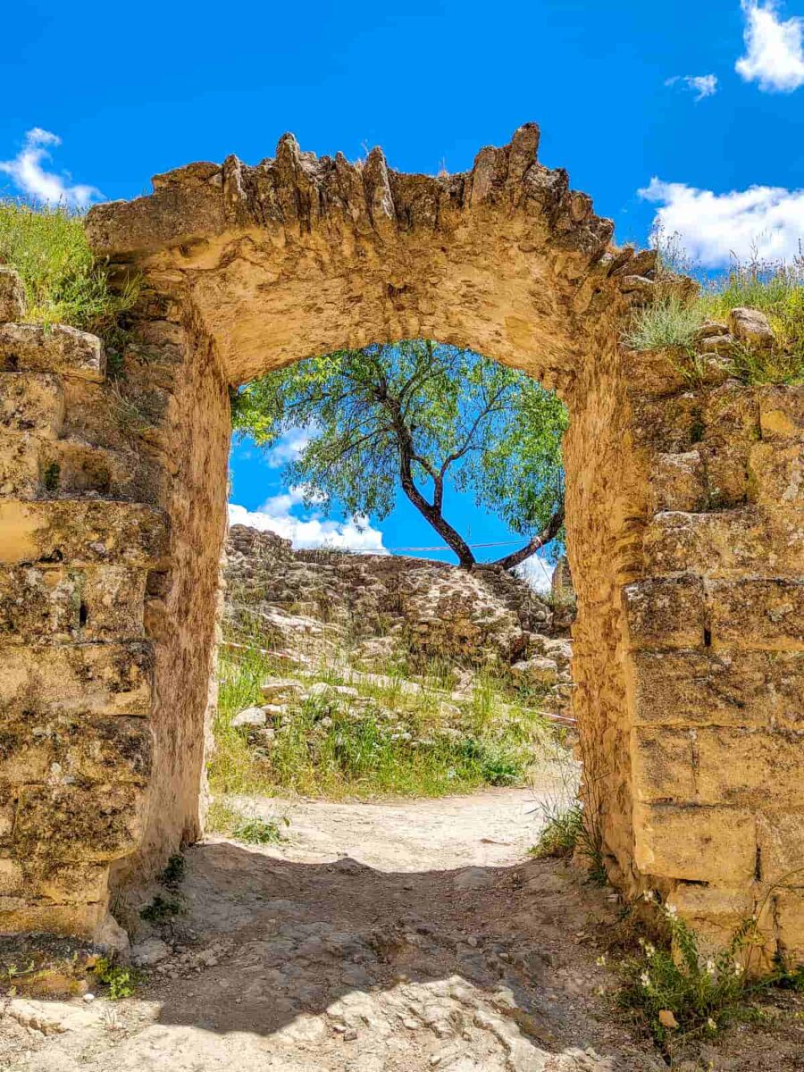 Stone archway ruins frame a view of a single tree and rocky terrain under a bright blue sky with scattered clouds. Grass and greenery are visible around the ancient stone structure. Remains of the Moorish fortress in Montefrío, Spain