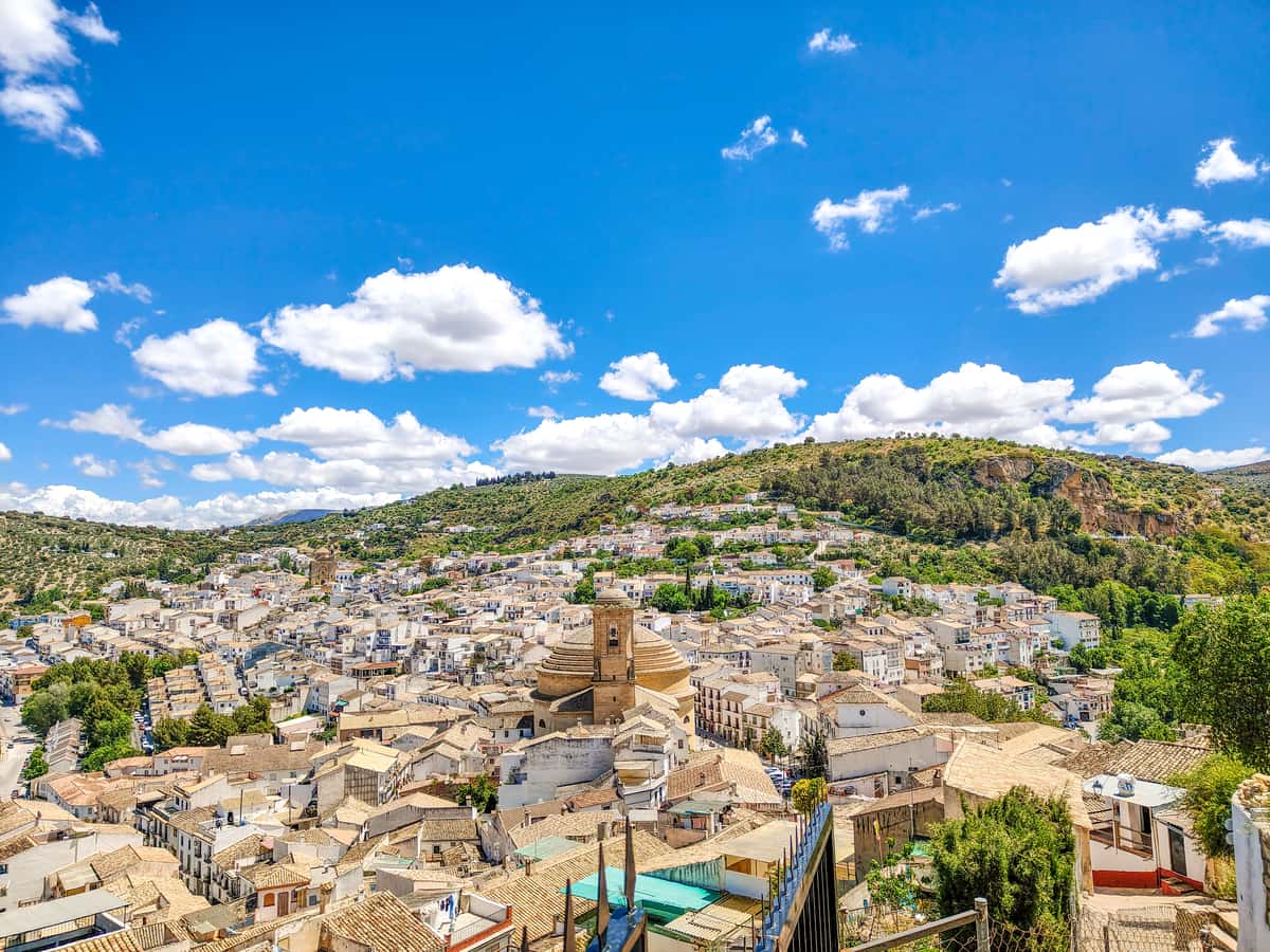 A panoramic view of a town with tan-colored buildings and a central church, nestled in a valley surrounded by green hills under a bright blue sky with scattered white clouds. View from Barrio del Arrabal in Montefrío, Granada