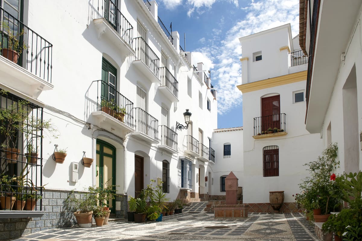 A sunlit narrow street with white buildings, balconies with black railings, potted plants, and patterned stone pavement. A small fountain stands in the center under a partly cloudy sky. A street in Cómpeta