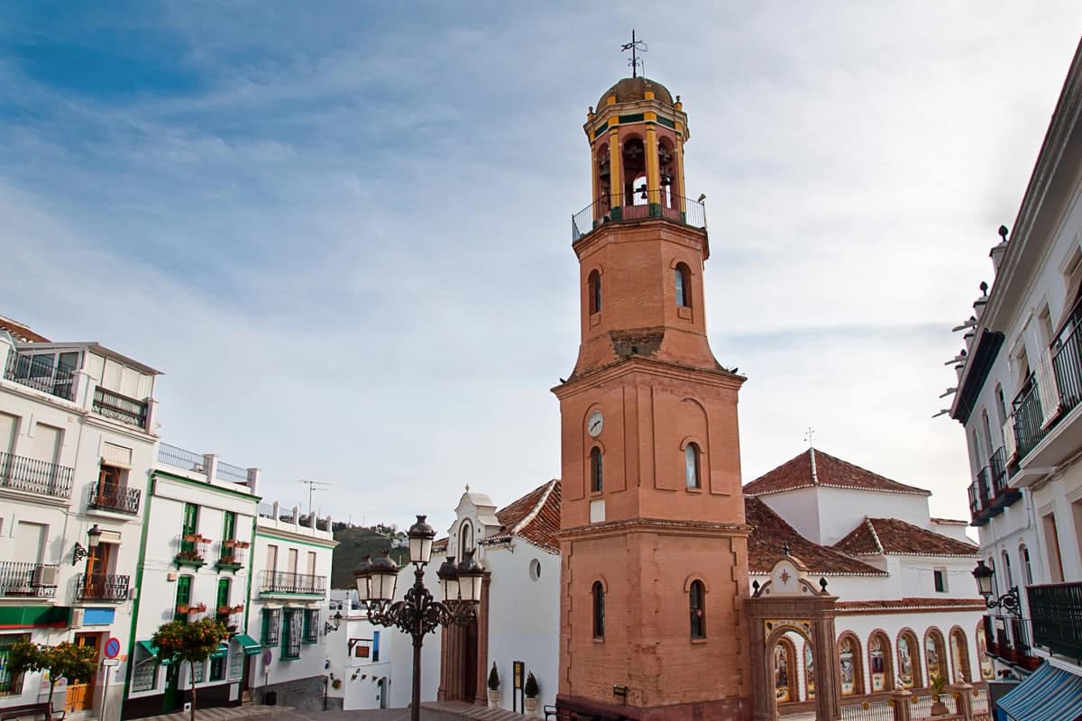 A tall, brown bell tower with arches stands in a town square surrounded by white buildings with balconies and traditional red-tiled roofs under a partly cloudy sky. Parroquia Nuestra Señora de la Asunción - one of the best things to do in Cómpeta