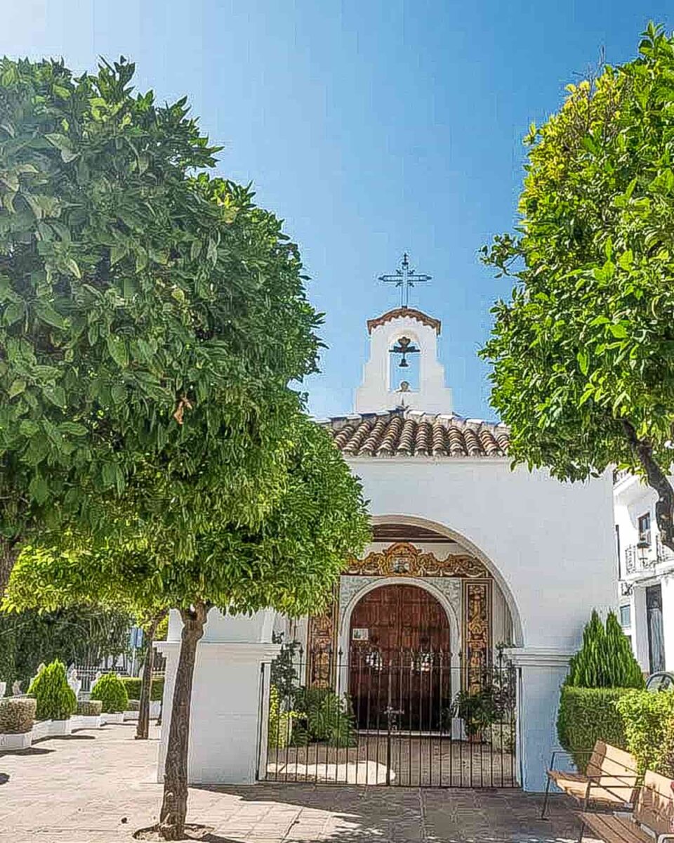 A small white church with a tiled roof and bell tower stands behind a gated entrance, flanked by two leafy green trees under a clear blue sky. A wooden bench is visible on the right side of the foreground. Ermita de San Antón in Cómpeta