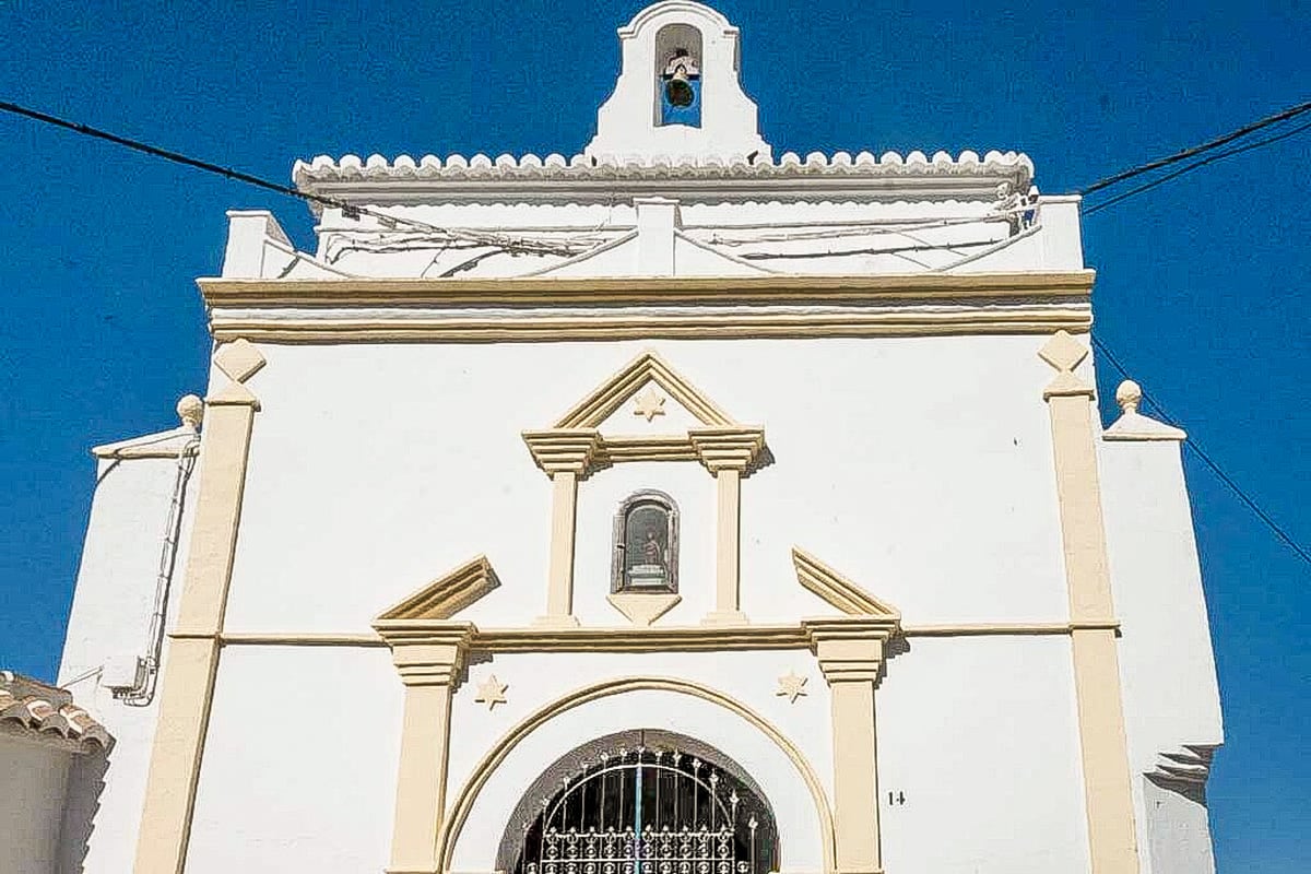 A white church façade with simple architectural details, a bell tower at the top center, and an arched doorway with a metal gate. The building stands against a clear blue sky. Ermita de San Sebastian in Cómpeta