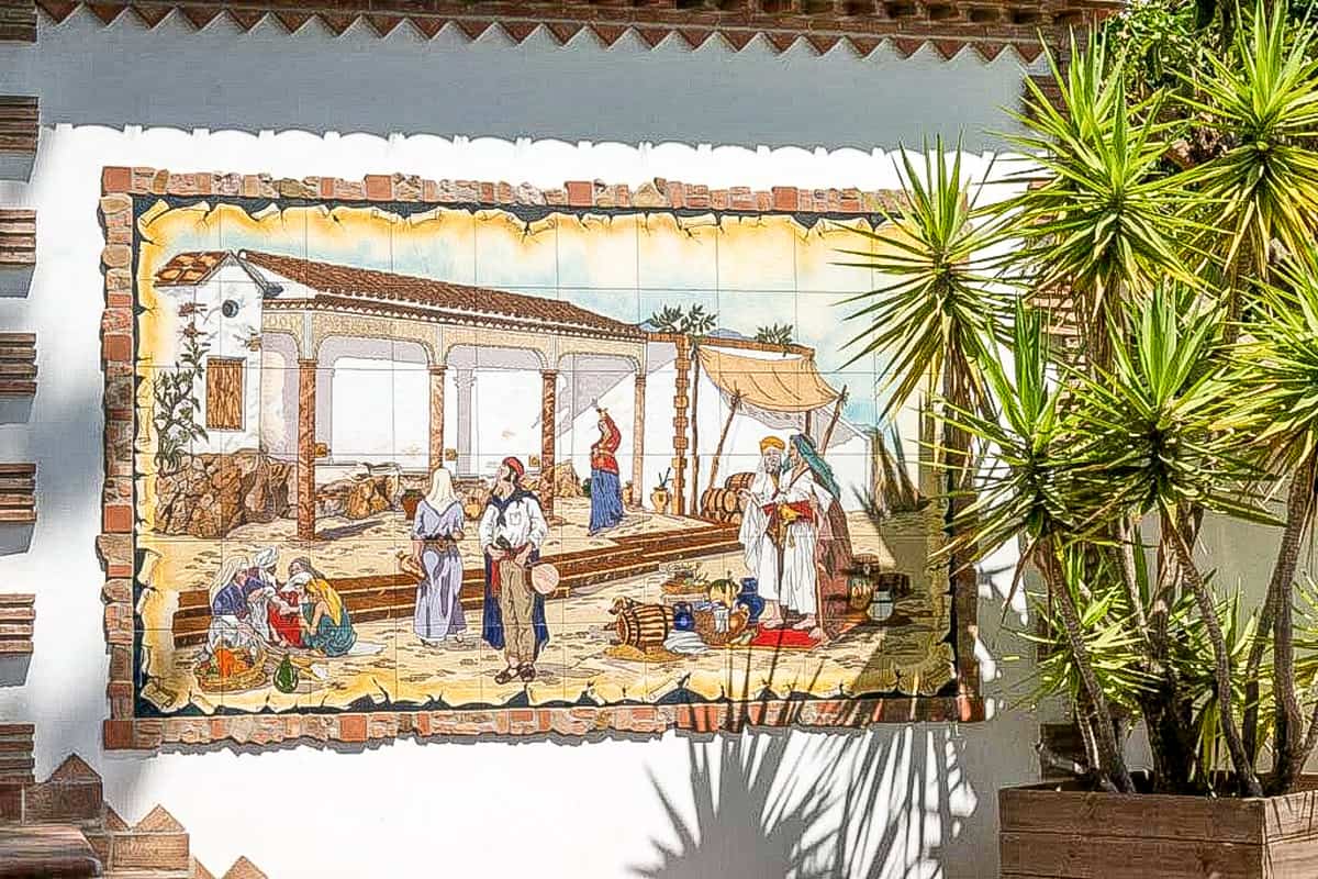 A colorful ceramic tile mural on an exterior wall depicts people in traditional clothing engaging in daily activities in a Mediterranean courtyard; potted palm plants are beside the mural. Mural in Cómpeta