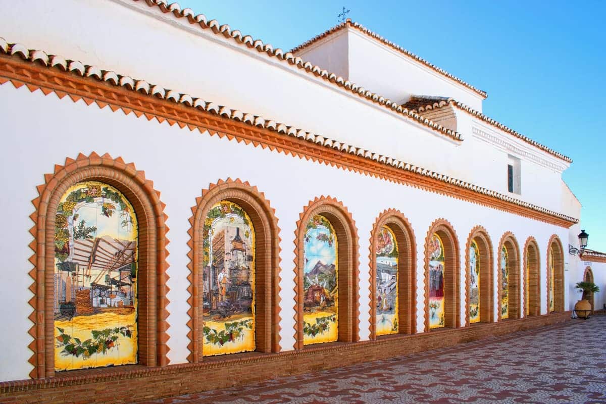 A white building with a row of arched windows framed by red bricks, each featuring colorful tile murals depicting various scenes; a brick-patterned walkway runs alongside the building. Paseo de Tradiciones in Cómpeta