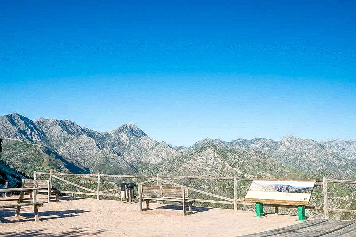A paved scenic overlook with wooden benches, a trash can, and a large information sign faces a view of rugged mountains under a clear blue sky. A wooden fence borders the viewpoint. Puerto Collardo Viewpoint near Cómpeta