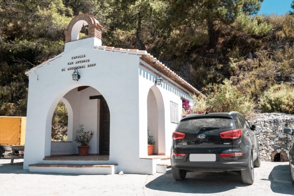 A small white chapel with an arched entrance and a bell above the door stands beside a black SUV parked in front. Trees and shrubs surround the building, and sunlight casts shadows on the ground. Capilla San Antonio in El Acebuchal