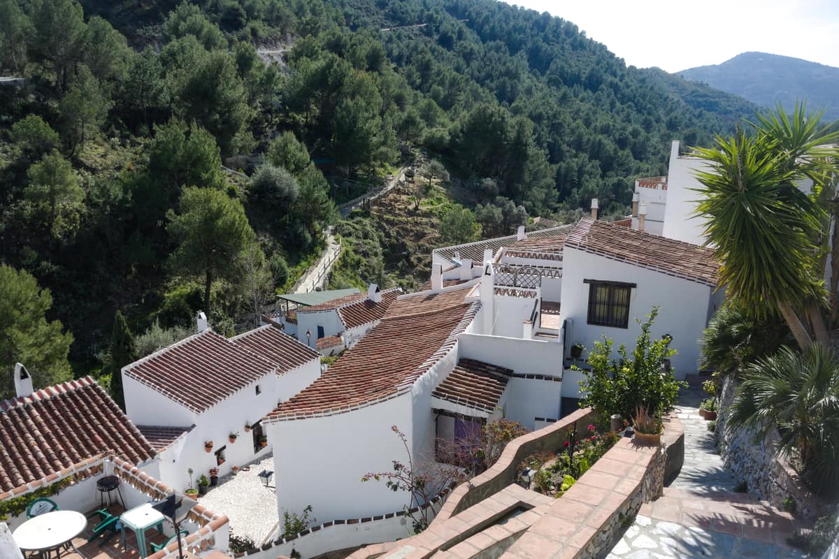 Whitewashed houses with red-tiled roofs are clustered on a slope, surrounded by green trees and mountains under a clear sky. A narrow path winds through the scene, bordered by plants and a stone wall. El Acebuchal near Frigiliana