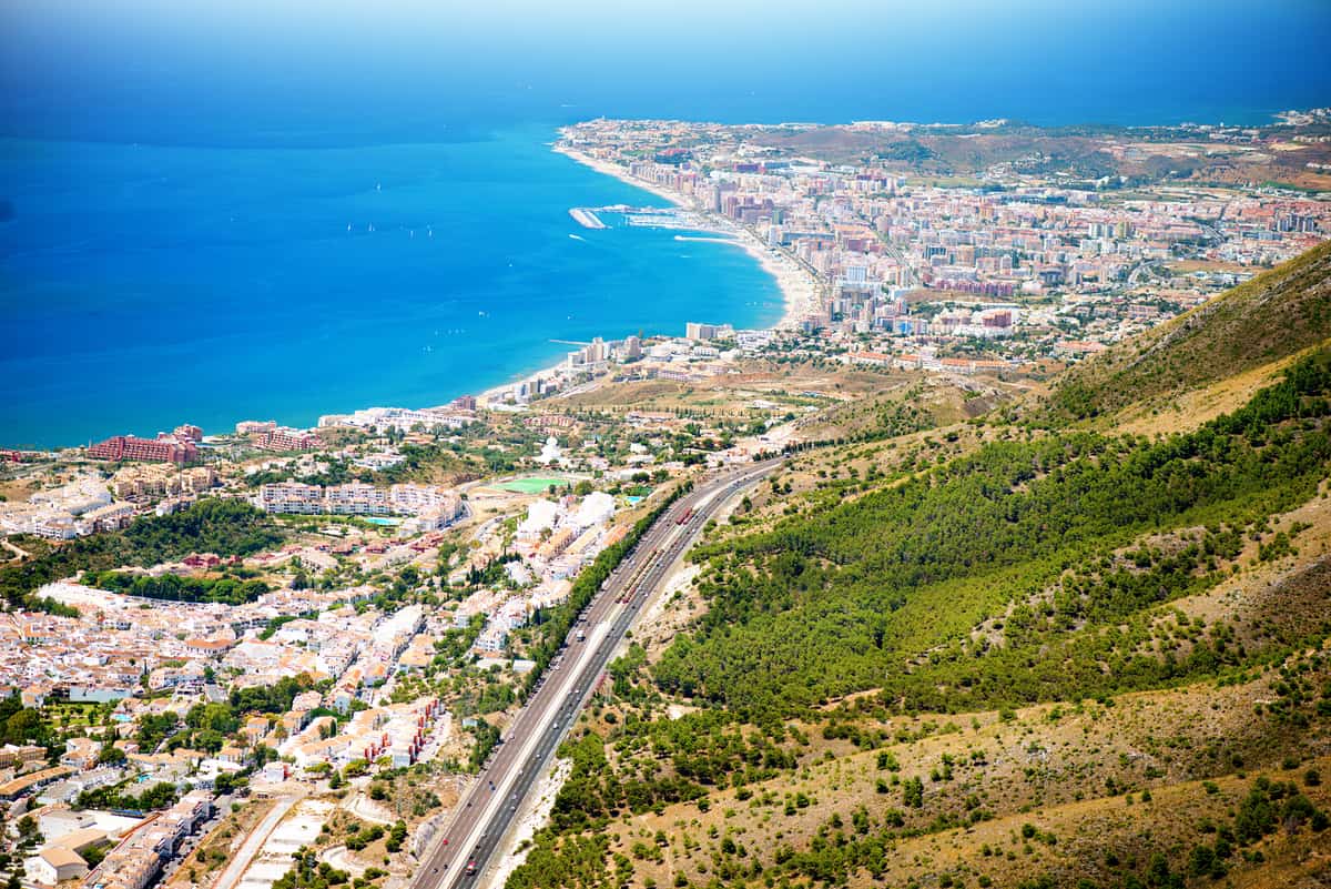 Aerial view of a coastal city with a long beach, turquoise sea, urban buildings, and green hills in the foreground under a clear blue sky. Costa del Sol, Andalsuai