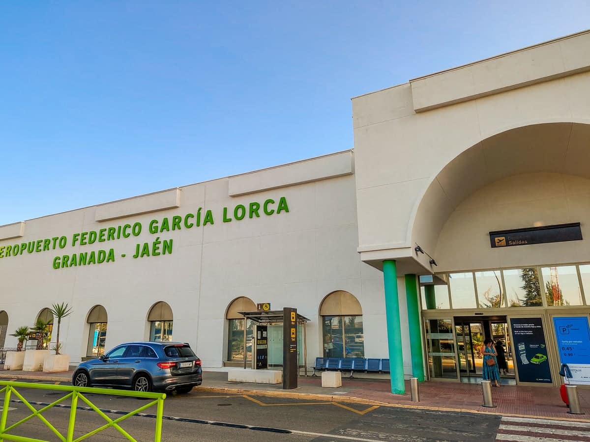 Exterior view of Aeropuerto Federico García Lorca Granada-Jaén, showing a white building with arched entrance, green signage, parked cars, and a person walking near the entrance on a sunny day.
