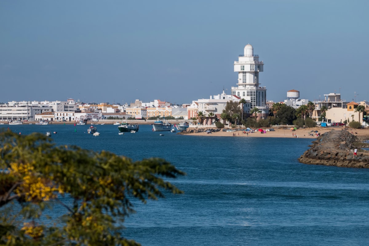A coastal town with white buildings, a prominent round lighthouse, and boats on blue water. People relax on the sandy beach. The scene is framed by greenery in the foreground under a clear blue sky. Isla Cristina, Huelva, Costa de la Luz