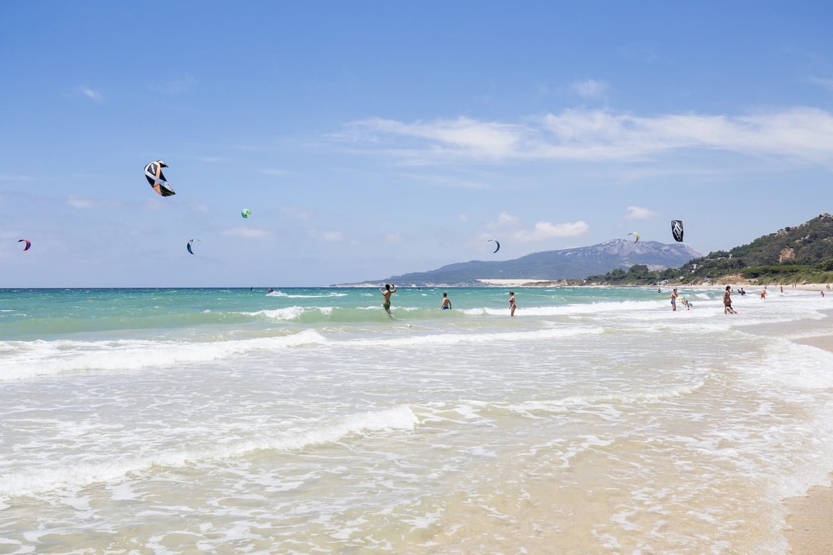People enjoy the beach and sea waves while several kite surfers ride the water under a clear blue sky with distant mountains and greenery in the background. Kite surfers in Tarifa, Costa de la Luz