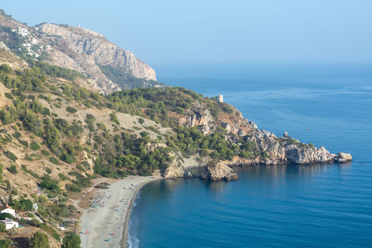A curved sandy beach bordered by clear blue sea and rocky cliffs covered with green trees, with mountains rising in the background under a clear sky. La Herradura, Granada, Costa Tropical