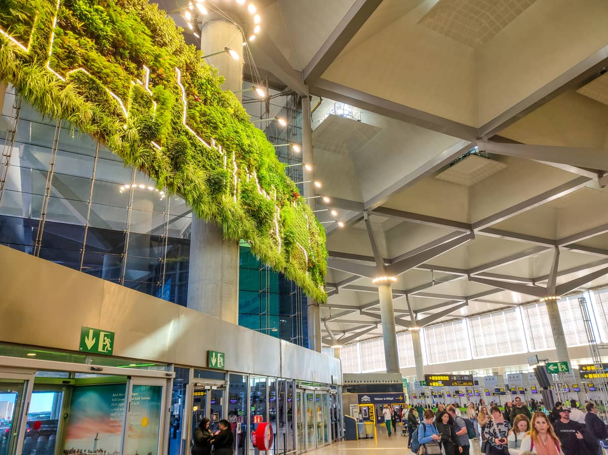 A modern airport terminal with a large vertical garden on the wall, glass doors, people walking with luggage, and bright overhead lighting. Inside Malaga Airport