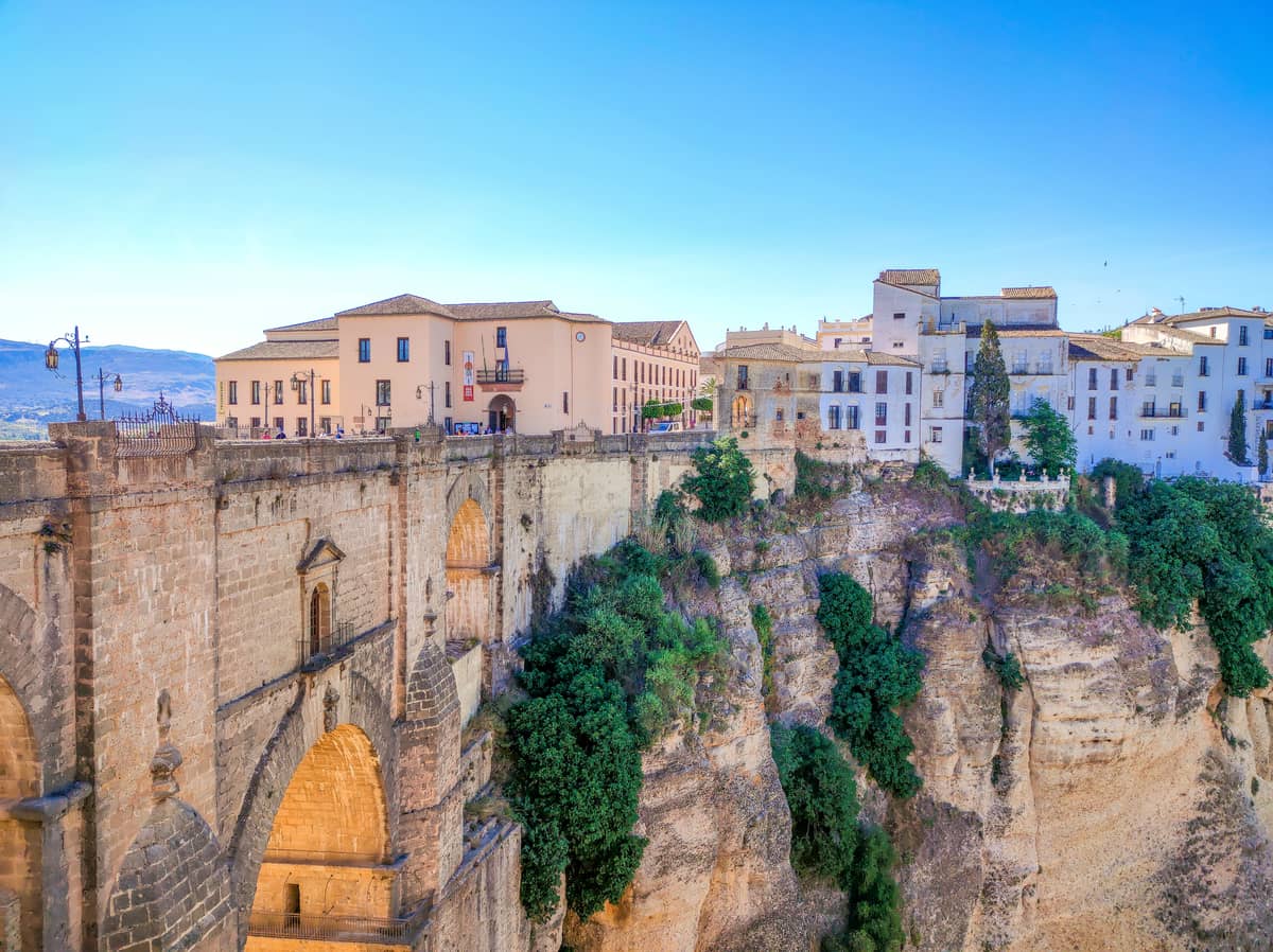 Historic stone bridge spans a deep gorge with white and tan buildings perched on the cliff edge in Ronda, Spain, under a clear blue sky.