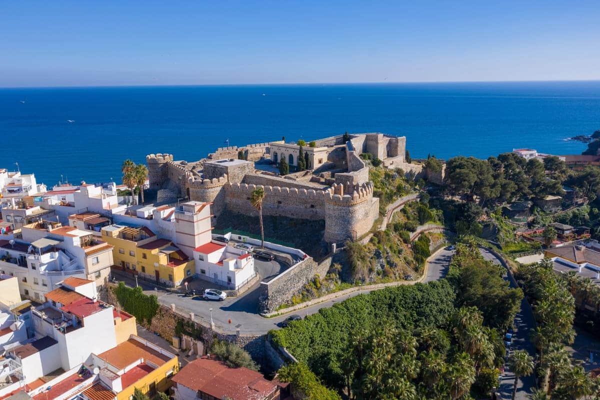 Aerial view of a large stone castle on a hill overlooking the sea, surrounded by colourful houses and lush greenery under a clear blue sky. Salobreña, Granada. Costa Tropical