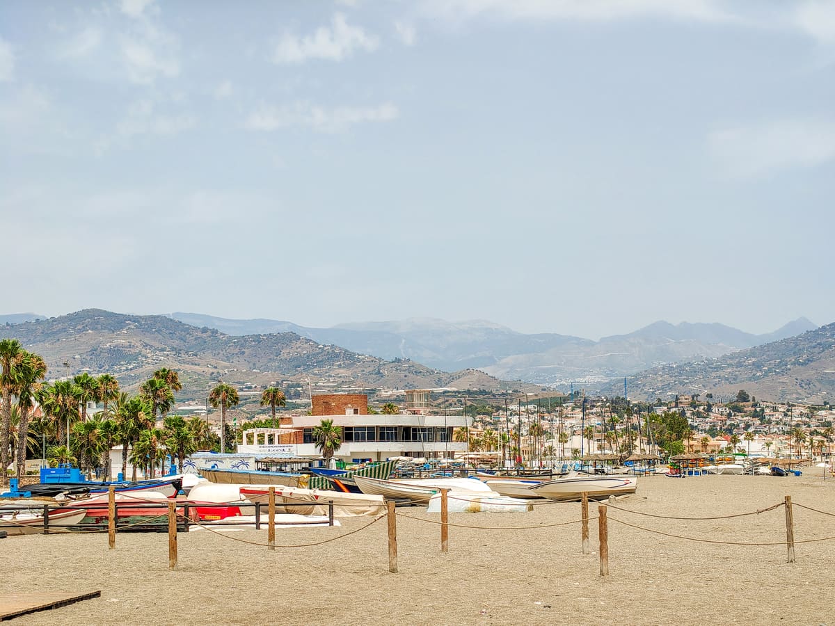 A sandy beach with small boats and kayaks on shore, palm trees, and buildings in the background. Hills and mountains rise in the distance under a clear, light blue sky. The beach of Torre del Mar on Costa del Sol