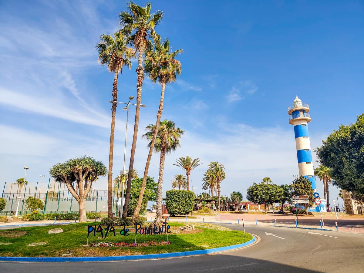 A sunny roundabout with tall palm trees, a sign reading "Playa de Poniente," and a blue-and-white striped lighthouse in the background against a clear blue sky. The lighthouse of Torre del Mar, Costa del Sol